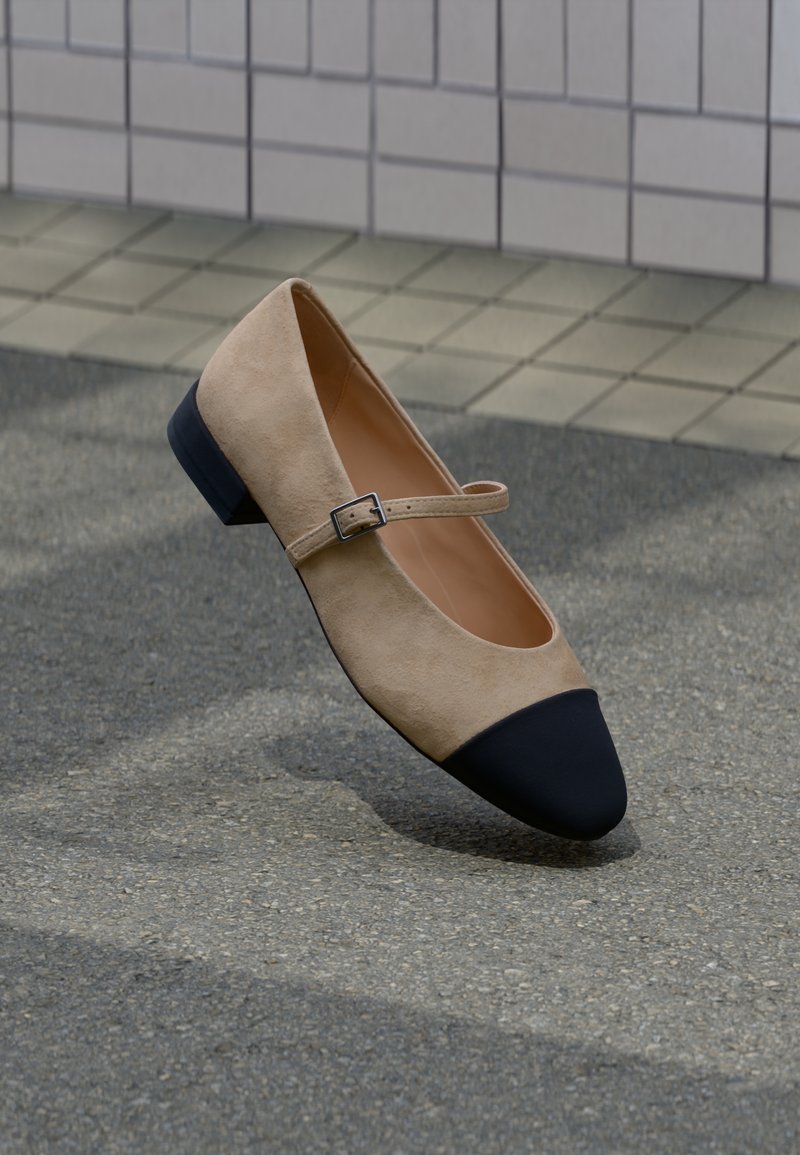 Chaussure Mary Jane en daim beige avec embout rond noir et petite bride à boucle, présentée sur une surface extérieure pavée.