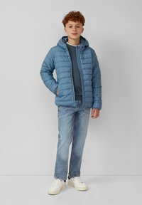 Blauer Pufferjacke mit Kapuze und gestepptem Design, getragen über einem dunklen Oberteil, kombiniert mit hellblauen Jeans und weißen Sneakern.