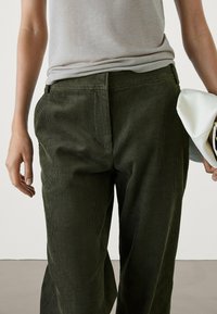 Pantalon en velours côtelé vert avec des rainures verticales, de coupe droite et poches latérales, associé à une chemise gris clair et une pochette blanche.