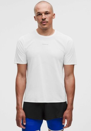 Mand med barberet hoved iført en hvid Lululemon t-shirt med korte ærmer, sorte shorts og blå kompressionstights, der står mod en ensfarvet baggrund.