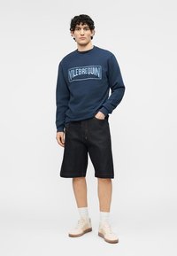 Homme portant un sweat-shirt bleu marine avec le texte « VILEBREQUIN », un short noir à hauteur des genoux, des chaussettes blanches et des baskets beiges, debout avec une main dans la poche.