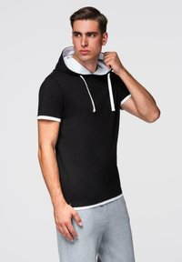 Mladý muž upravuje kapuci na svém černém tričku s krátkým rukávem s bílými detaily, nosí světle šedé kalhoty, na jednobarevném pozadí.