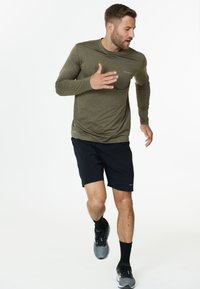Olivgrünes Langarm-Sportshirt, kombiniert mit schwarzen Shorts. Das Shirt hat eine enganliegende Passform und beide Kleidungsstücke bestehen aus leichtem Stoff.