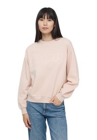 Jasnoróżowy sweter z haftem "AMOUR" w kursywie. Posiada okrągły dekolt, długie rękawy i luźny krój. Noszony z niebieskimi dżinsami.