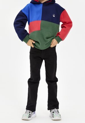 Kind trägt einen Kapuzenpullover mit Farbblockdesign in Blau, Navy, Rot und Grün, schwarze Hose und weiße Turnschuhe, steht mit Händen in den Vorder Taschen.