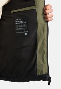 Black and olive green jacket interior with a smooth texture, featuring a care label, pocket, and zipper with a branded pull tab.