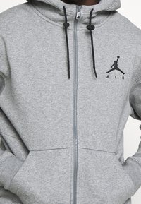 Szara bluza zapinana na zamek, wykonana z miękkiego materiału, z czarnym logo Jumpman na klatce piersiowej, regulowanym kapturem z troczkami oraz przednimi kieszeniami.