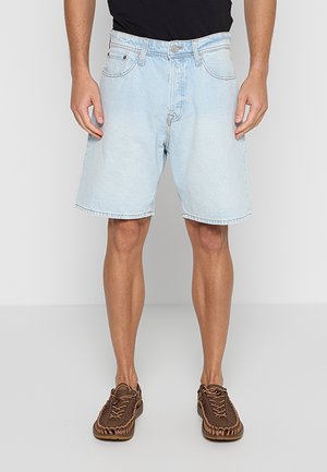 Mężczyzna ubrany w jasnoniebieskie szorty jeansowe, czarną koszulę i brązowe plecione buty sznurowane, stoi na tle jednolitego jasnoszarego tła.
