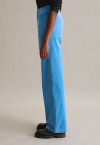 Pantalon large en velours côtelé bleu avec une finition texturée, comprenant des poches latérales et une taille haute, associé à des chaussures à plateforme noires.