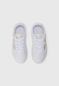 Hvite sneakers med lav topp, hvite lisser og gull "EA7 Emporio Armani" logo på tunger og sider, vist side om side på hvit bakgrunn.