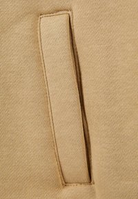 Beige stof med en glat tekstur, der har en lodret, syet lomme. Sømmen er pæn, og stoffet viser et fint strikkemønster.