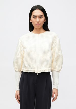 Kort, cremefarvet jakke i tekstureret stof med lynlås foran og rynkede manchetter. Kombineret med et mørkt outfit og lange ærmer.