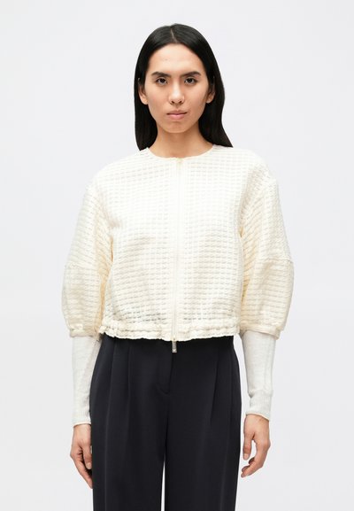 Veste courte de couleur crème en tissu texturé avec fermeture éclair à l'avant et poignets froncés. Associée à une tenue foncée et des manches longues.