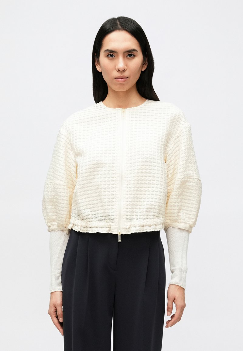 Κοντό, εκρού μπουφάν από υφασμάτινη υφή με εμπρός φερμουάρ και σούρες στα μανίκια. Συνδυάζεται με σκούρο σύνολο και μακριά μανίκια.