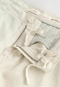 Pantalones cortos de lino beige claro con cierre de cremallera y botón, que cuentan con un forro interior a rayas y cordón ajustable, mostrando una tela suave y texturizada.