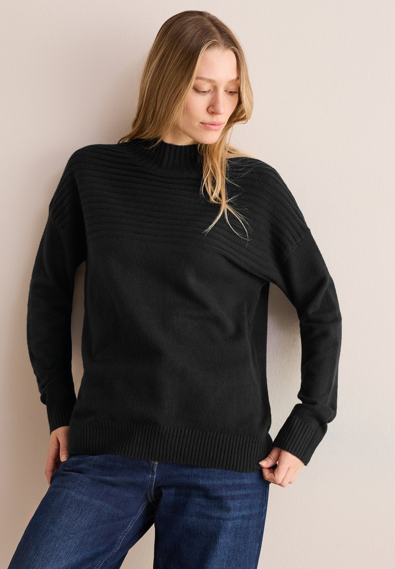 Maglione nero con un colletto alto a costine, caratterizzato da righe orizzontali testurizzate nella parte superiore, e polsini e fondo a costine. Indossato con jeans blu.