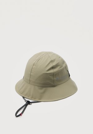 Sombrero tipo pescador de ala ancha color beige claro con correa negra ajustable para la barbilla y logo sutil en el lateral, mostrado sobre un fondo liso.