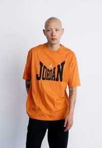 Orange bomulds T-shirt med et sort "JORDAN" logo og grafisk motiv ovenfor. Løs pasform med korte ærmer og rund hals.