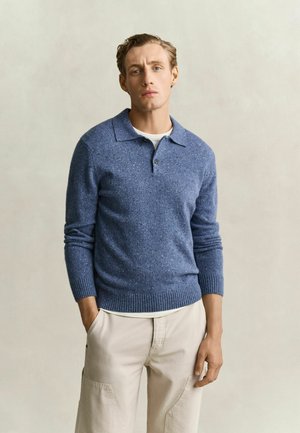 Man met lichtbruin haar, gekleed in een blauw gebreide poloshirt met lange mouwen en beige broek, staat met zijn handen gedeeltelijk in zijn zakken.