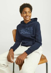 Temno modra kapuca s logotipom "JACK & JONES", v kombinaciji z svetlo sivo trenirko. Model sedi na leseni stolu.