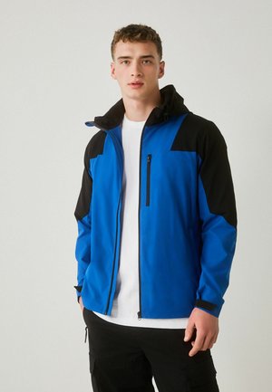 Young man wearing a blue and black zip-up jacket with hood, white t-shirt, and black cargo pants against a plain light background.