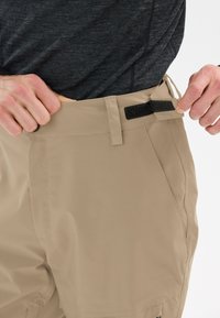 Pantalon beige au toucher lisse, avec une fermeture Velcro à la taille et de grandes poches latérales. Le tissu semble léger et durable.