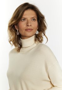 Beige turtleneck sweater with a soft texture, featuring ribbed accents at the neck. Model also wears round, patterned earrings.