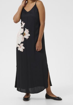 Femme portant une longue robe noire sans manches avec un grand imprimé floral rose pâle et des fentes latérales, associée à des chaussures plates noires.