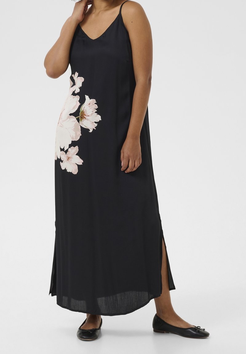 Femme portant une longue robe noire sans manches avec un grand imprimé floral rose pâle et des fentes latérales, associée à des chaussures plates noires.