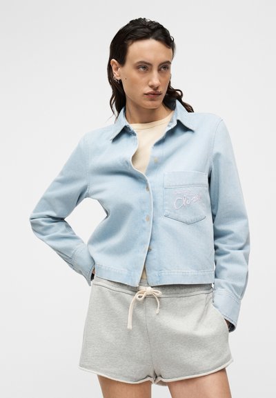 Ανοιχτό μπλε τζιν μπουφάν με κούμπωμα με κουμπιά και μπροστινή τσέπη. Διακοσμημένο με το κέντημα «Classic» σε ροζ χρώμα. Συνδυασμένο με γκρι σορτς.