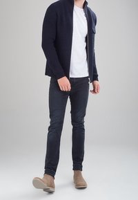 Granatowy prążkowany kardigan z pełnym zamkiem, noszony na białej koszulce i zestawiony z ciemnymi dżinsami oraz beżowymi botkami za kostkę.