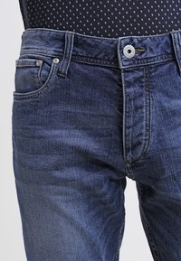 Mörkblå denimjeans med en slät yta, featuring dragkedja och knappstängning, femficksdesign och lätta blekningar.