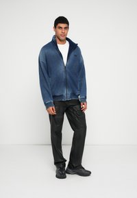 Modra jakna z zadrgo in teksturirano površino, nošena preko bele majice, kombinirana s črnimi usnjenimi hlačami z žepi in črnimi čevlji.