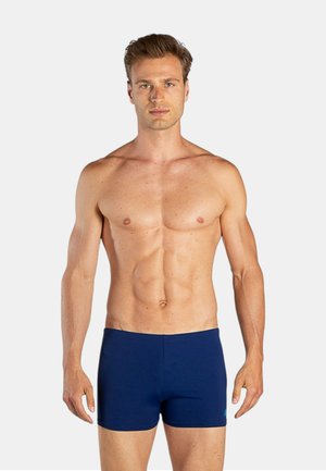 Uomo a torso nudo con capelli corti sta in piedi rivolto in avanti, indossa un costume da bagno aderente blu navy su sfondo chiaro e uniforme.