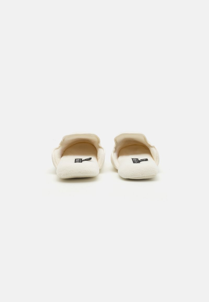 Chaussons ivoire duveteux avec un bout arrondi, une texture douce et un design minimaliste. Accent de label noir sur le côté intérieur pour le branding.