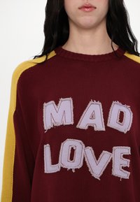 Maroon knit sweater featuring "MAD LOVE" in frayed pastel appliqué. Accented with yellow shoulder panels and a ribbed neckline.