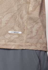 Beżowy top sportowy z teksturowanym wzorem kropek i kształtów, z małą metką ACTIBREEZE i przeszyciami. W połączeniu z szarymi szortami.