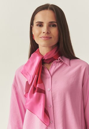 Moteris dėvi šviesiai rožinę marškinėlių su sagutėmis, apie kaklą surištą rožinę ir raudoną raštuotą šilkinį šaliką bei derančias auskarus.