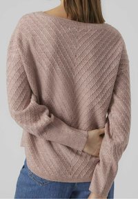 Mujer vistiendo un suéter de punto trenzado rosa con un diseño en V, jeans azules y la mano izquierda metida detrás de la espalda.