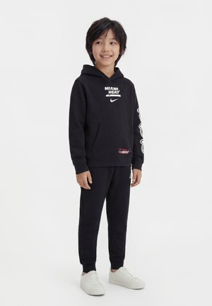 Bambino sorridente in piedi, che indossa una felpa con cappuccio nera dei Miami Heat e pantaloni abbinati con scarpe da ginnastica bianche, su sfondo semplice.