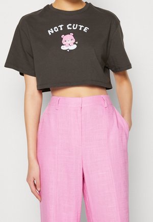 Persoon draagt een donker kort T-shirt met de tekst "NOT CUTE" en een roze berenillustratie, gecombineerd met felroze hoge taille broek.