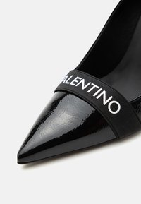 Chaussure noire brillante à bout pointu avec une bride élastique noire à l'avant affichant le mot « VALENTINO » en lettres blanches.