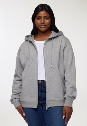 Donna che indossa una felpa grigia chiara con zip su una maglietta bianca e jeans blu, in piedi su uno sfondo bianco semplice.