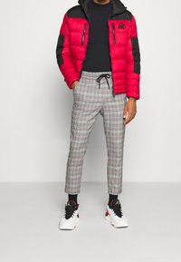 Röd och svart pufferjacka med dragkedjefickor, svart långärmad tröja, grå rutiga byxor och svarta sneakers med färgglada accenter.