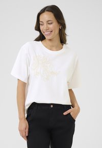 T-shirt blanc à manches courtes, avec une broderie florale beige sur le devant. Associé à un pantalon noir, offrant une coupe décontractée.