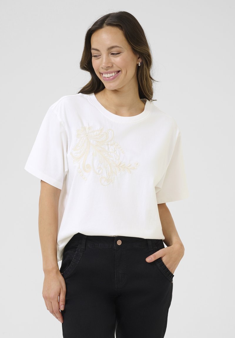 T-shirt blanc à manches courtes, avec une broderie florale beige sur le devant. Associé à un pantalon noir, offrant une coupe décontractée.