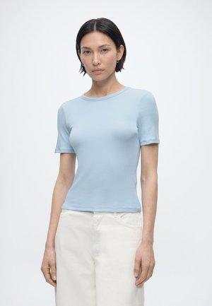 Femme aux cheveux courts et foncés portant une chemise à manches courtes bleu clair et un pantalon blanc, debout devant un fond blanc uni.