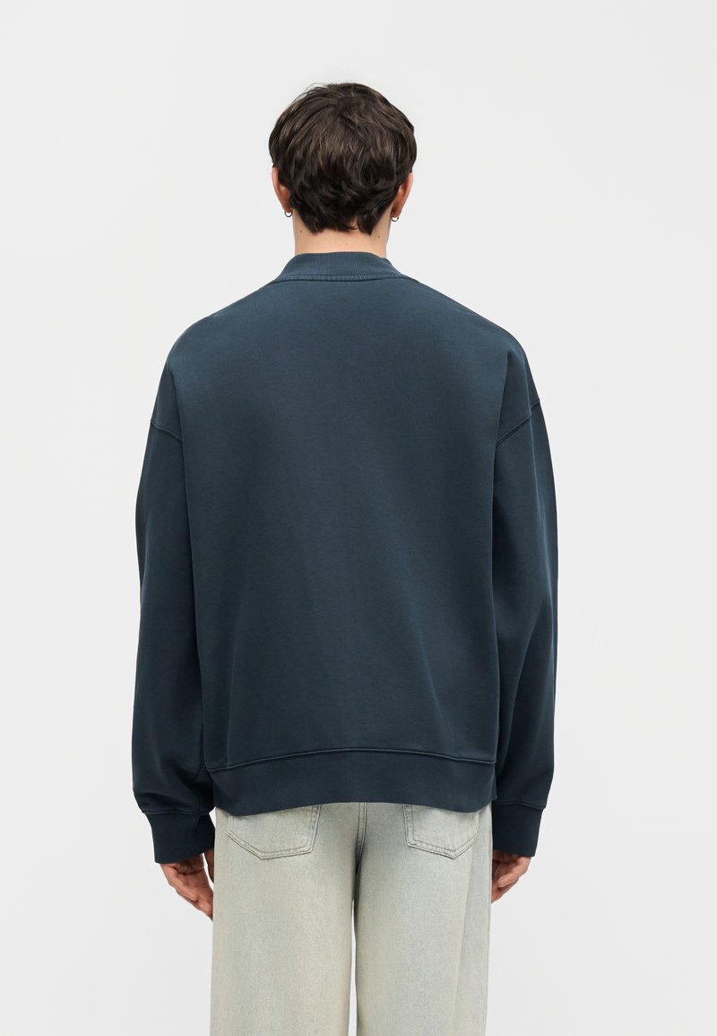 Man with short dark hair wearing a dark blue sweatshirt and light gray jeans, facing away against a plain light background.