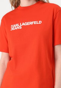 Rdeča bombažna majica s kratkimi rokavi, na kateri je v velikih belih črkah napisan "KARL LAGERFELD JEANS" prek prsi. Klasičen okrogli izrez in kratki rokavi.