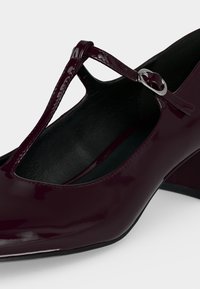 Chaussure Mary Jane en cuir verni bordeaux foncé avec bride en T et boucle argentée, talon bas bloc et doublure intérieure noire.